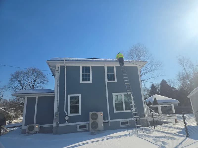 Person on a ladder removing snow from the roof edge of a two-story house