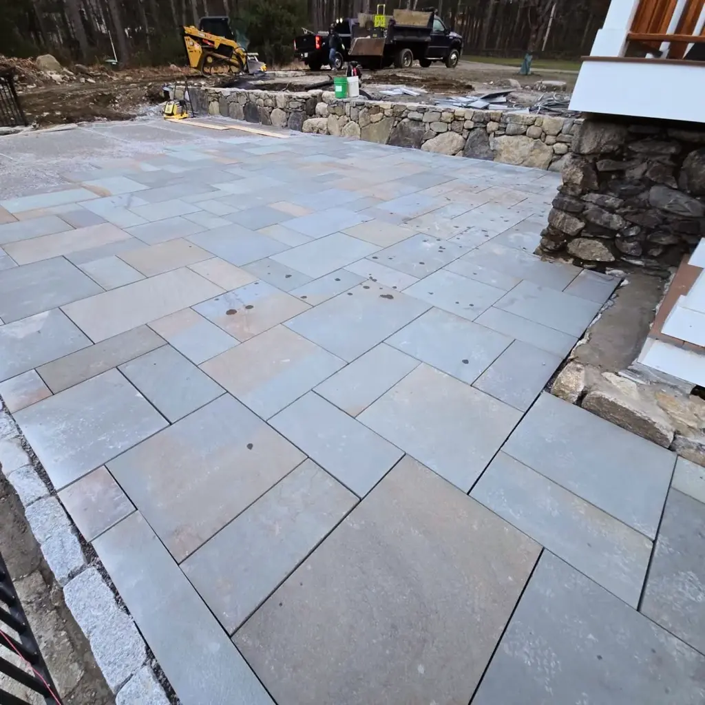 Square and rectangular gray stone pavers beside a low rock wall and a house foundation
