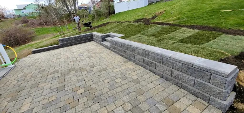 Paver patio beside gray block retaining walls and rows of sod on a sloped yard