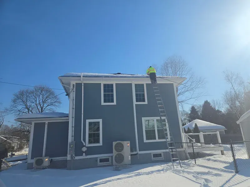 Person standing at the top of a ladder beside the snow-covered edge of a two-story house roof